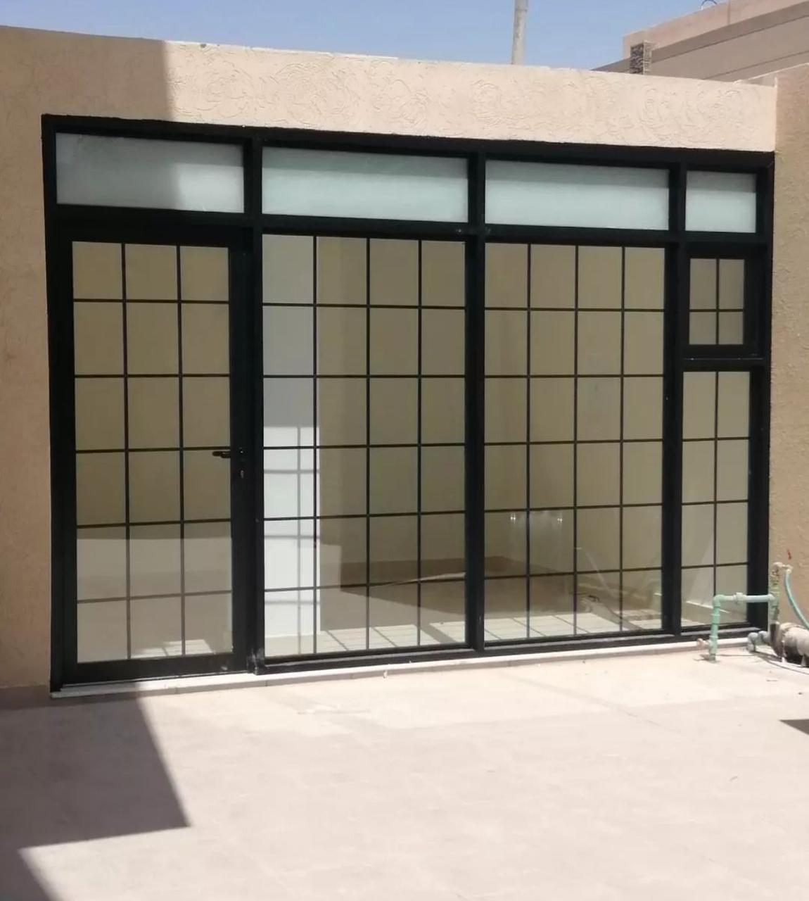 واجهة زجاجية كبيرة بإطار ألمنيوم أسود وتقسيمات مربعة، تتوسطها باب زجاجي مفصلي، مركبة في جدار خارجي مع فناء إسمنتي أمامها.