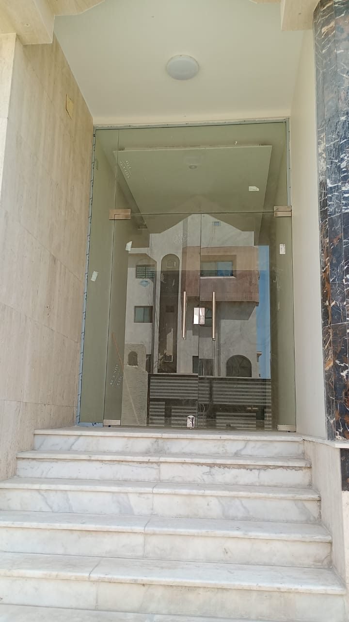 مدخل مبنى بباب من زجاج السيكوريت الشفاف بدون إطار مع قواطع زجاجية جانبية وملحقات تثبيت معدنية، يعلو درجًا رخاميًا بين جدارين حجريين.
