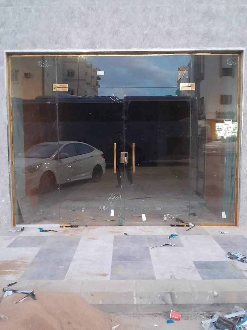 واجهة محل تجاري مركّبة بزجاج سيكوريت داكن ضمن إطار معدني رفيع، يتوسطها باب زجاجي مزدوج بمقابض، وأمامها رصيف عليه بقايا أعمال التركيب.