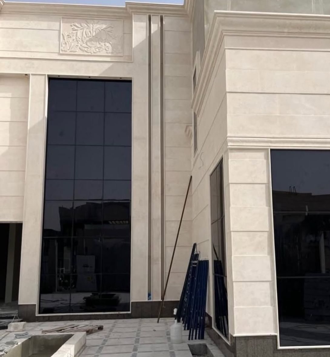 مبنى بواجهة حجرية فاتحة اللون تتوسطها واجهة زجاجية داكنة كبيرة بإطار معدني، وأمامها بقايا مواد بناء وعمود طويل مسند على الجدار.