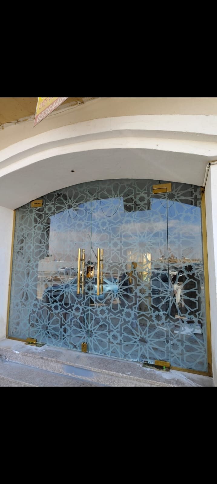 واجهة محل من الزجاج المقسى مع باب مزدوج بمقابض نحاسية ونقوش هندسية مزخرفة على الزجاج، ضمن إطار مقوس أبيض.