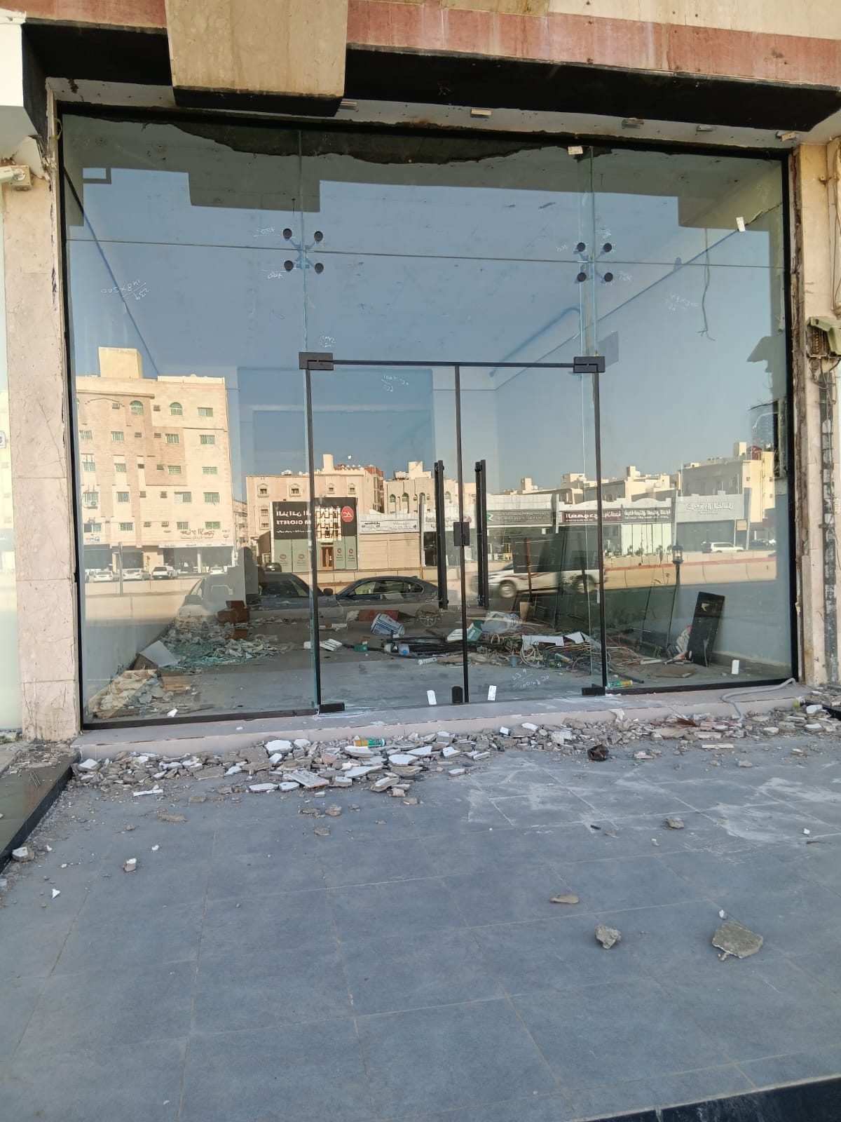 واجهة محل تجاري مركّبة بالكامل من زجاج سيكوريت شفاف، في وسطها باب زجاجي مزدوج بمقابض ستيل عمودية وقواعد تثبيت علوية وسفلية، ويظهر خلف الزجاج شارع ومبانٍ، مع بقايا أعمال بناء على الأرض.