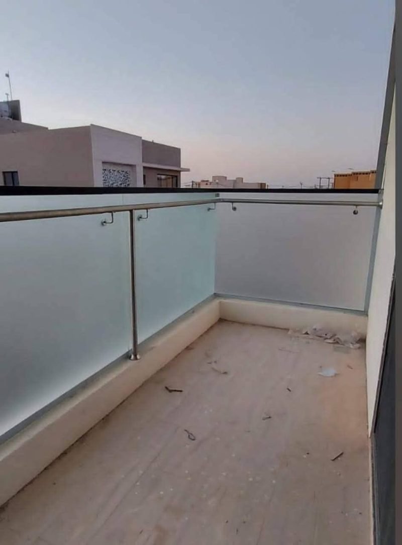 درابزين شرفة خارجية من ألواح زجاج سيكوريت مصنفر مثبتة بقواعد ومسكة من الستيل، مع أرضية شرفة غير مشطبة وأفق المدينة عند الغروب.