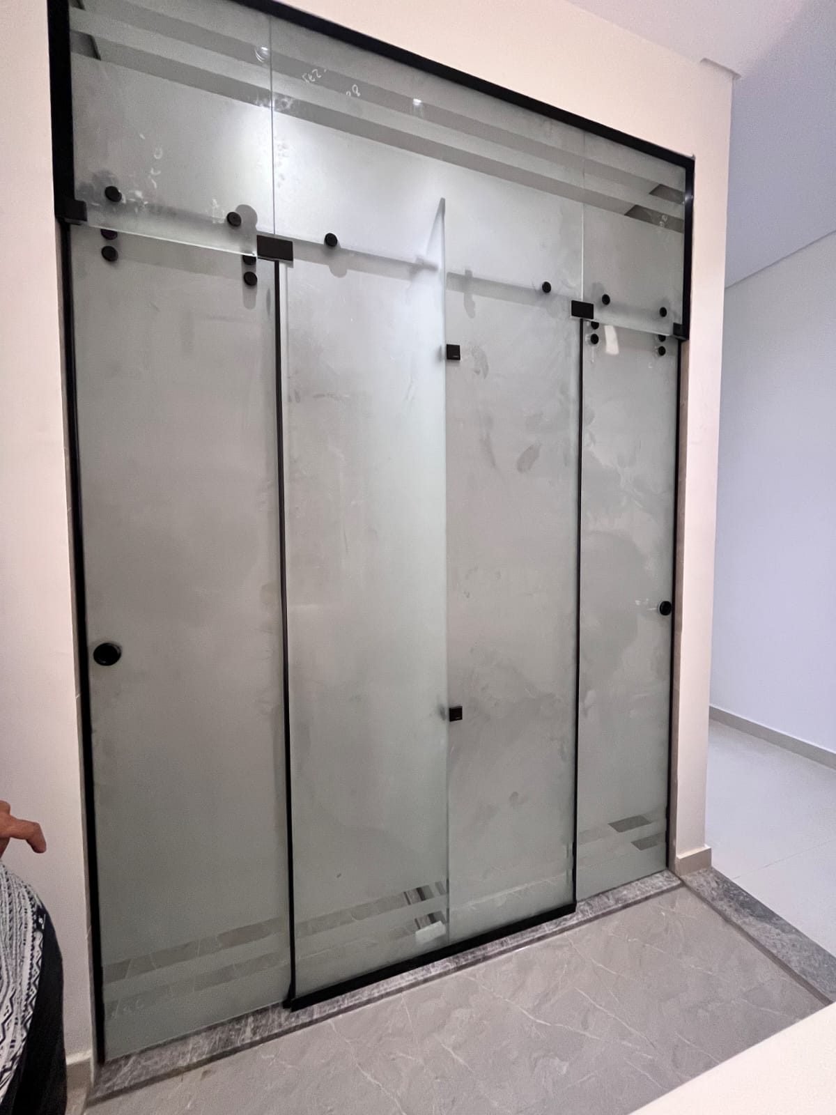 قاطع زجاجي شفاف من السيكوريت مكوّن من ثلاثة ألواح بإطار وإكسسوارات سوداء وباب مفصلي، يفصل منطقة الاستحمام في حمّام مبلّط برخام رمادي مع مصرف أرضي طولي.